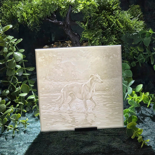 A square 3D printed plaque featuring an embossed dog walking through shallow water, displayed on a small stand in front of a green bonsai tree and foliage.