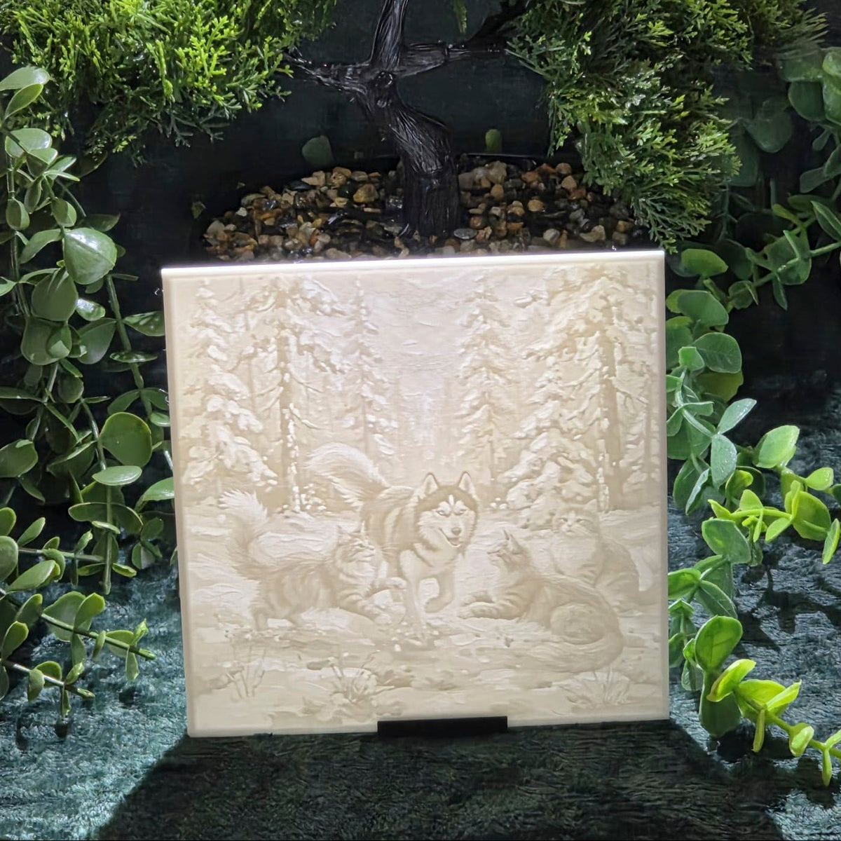 A square cream-colored 3D printed plaque featuring an embossed carving of a forest scene with cats and a dog, displayed in front of a faux bonsai tree and green foliage.